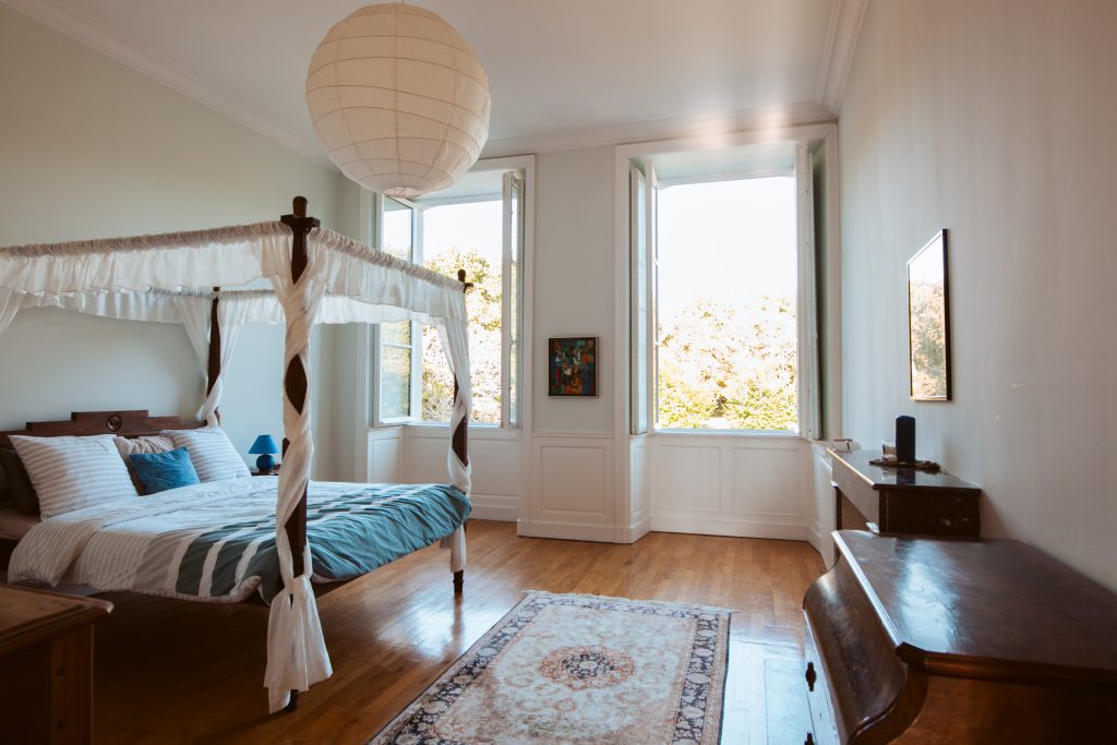 a spacious room with wooden parquet and a wooden frames double four poster bed. two bright windows are in the background