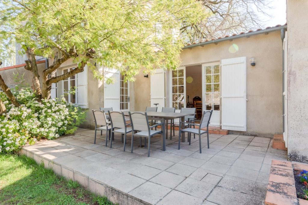 a patio in front of a house with white shutters with a n outdoor dining table and 8 chaires under a mimosa tree