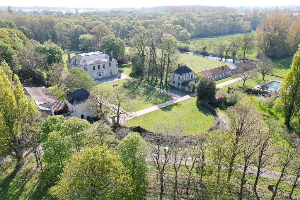 an aerial view of a property with large castle styled like a manor, a green front lawn with a low semi circle wall in front and some trees. To the sides there are other houses