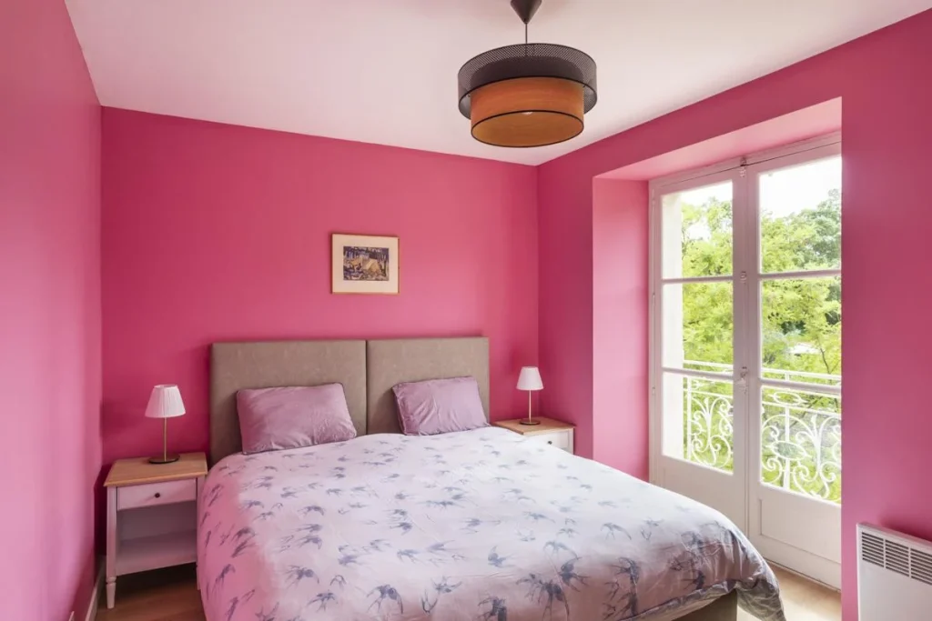 a bright room with pink walls and a double bed. On the side there is a French window and the bed has a bedside table and lamp on each side